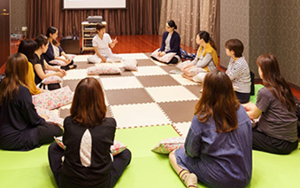 妊婦さんと助産師が輪になって床に座り、赤ちゃんのお世話について講習を受けている場面の写真です。